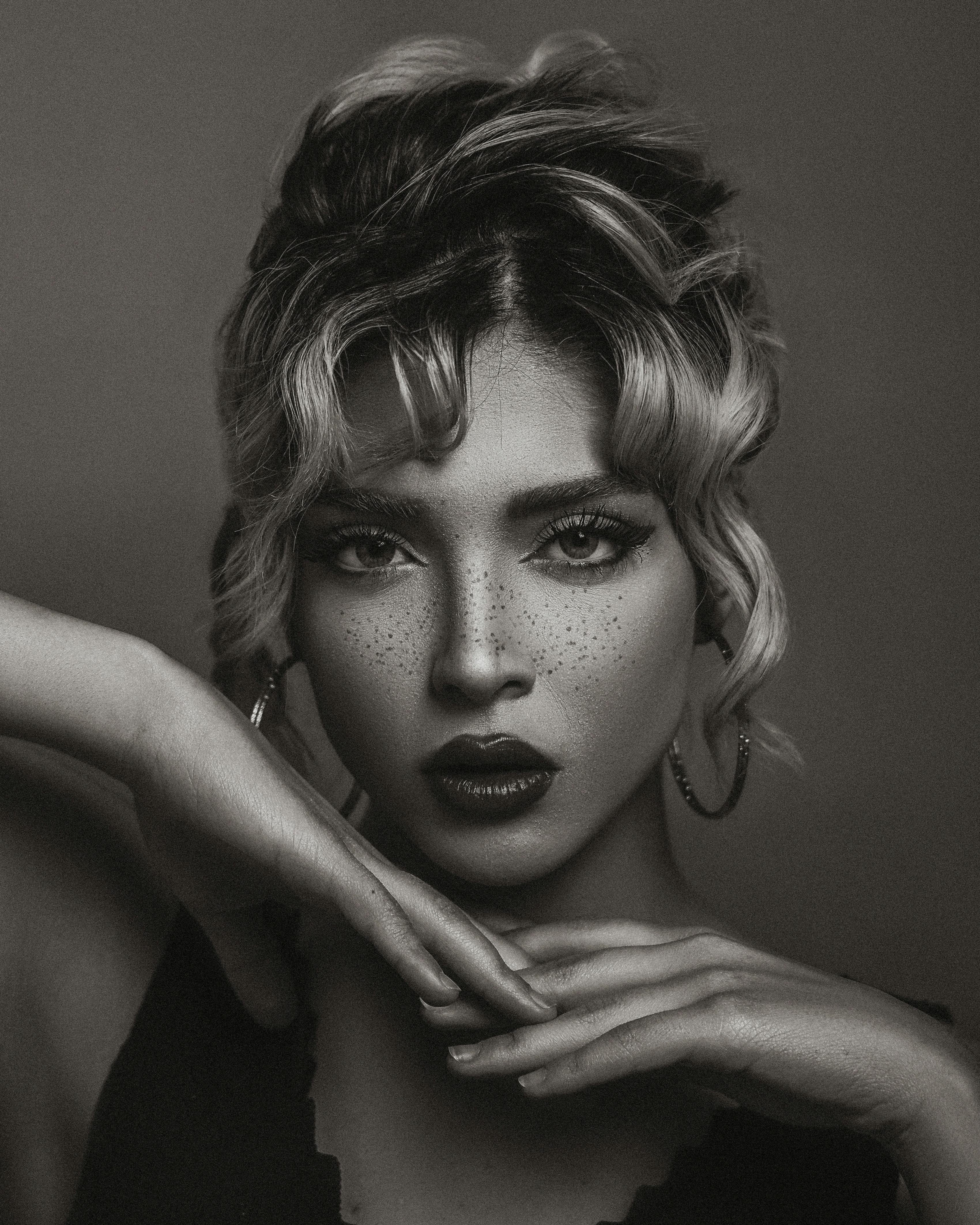 Black and white portrait of a blonde woman with hands under chin in a dramatic pose.