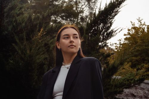 Free Young woman in a black jacket posing stylishly outdoors with greenery. Stock Photo