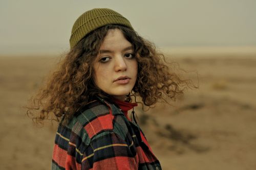 Free A young woman with curly hair wearing a plaid shirt and beanie stands outdoors with a thoughtful expression. Stock Photo