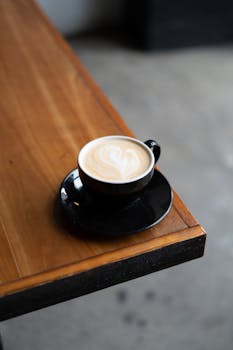 A warm cappuccino with latte art in a black cup, sitting on a wooden table indoors.