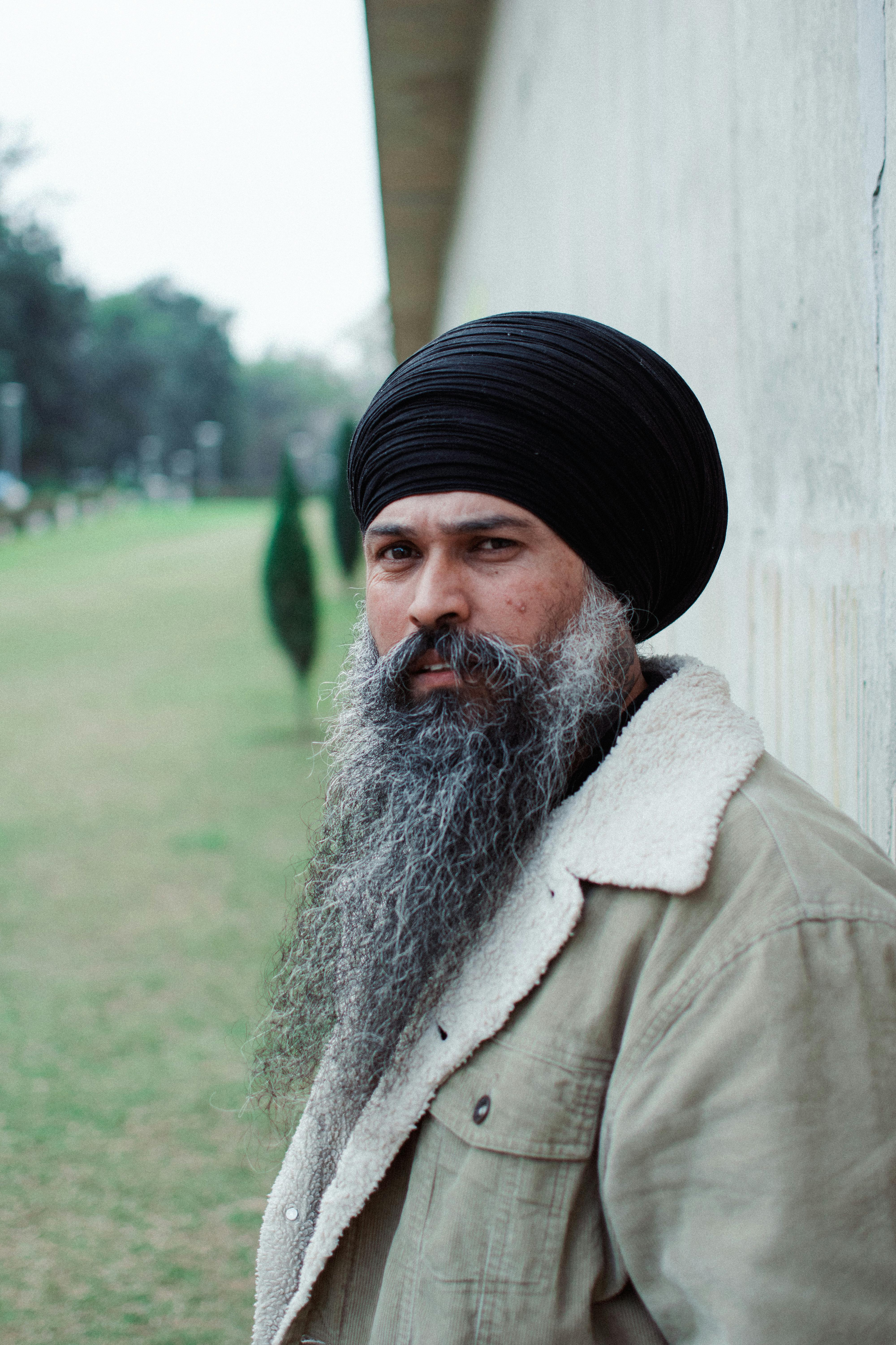 Side view portrait of a man with a turban and long beard standing by a wall in a park setting.