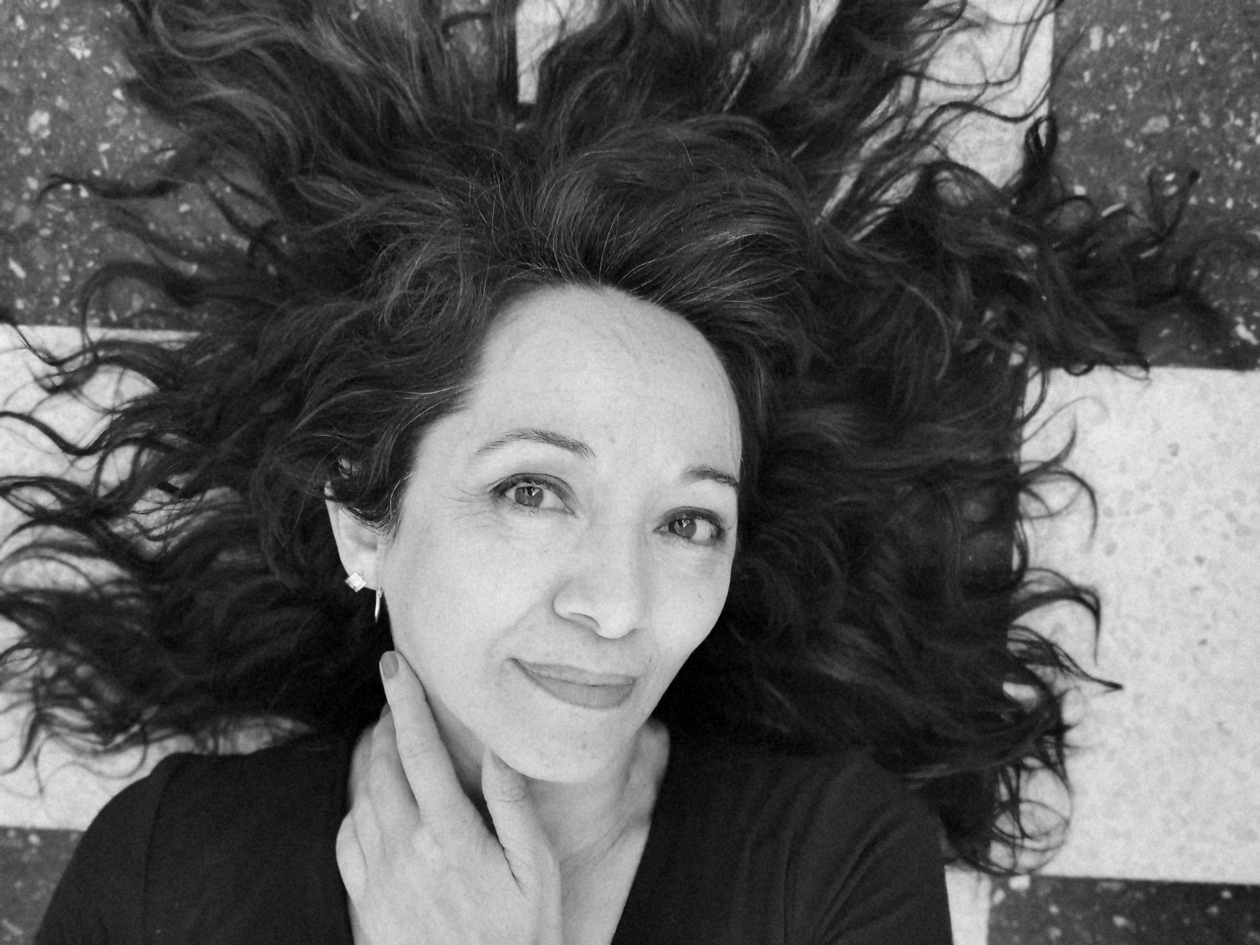 A close-up black and white portrait of a smiling woman with long hair lying on a patterned surface.
