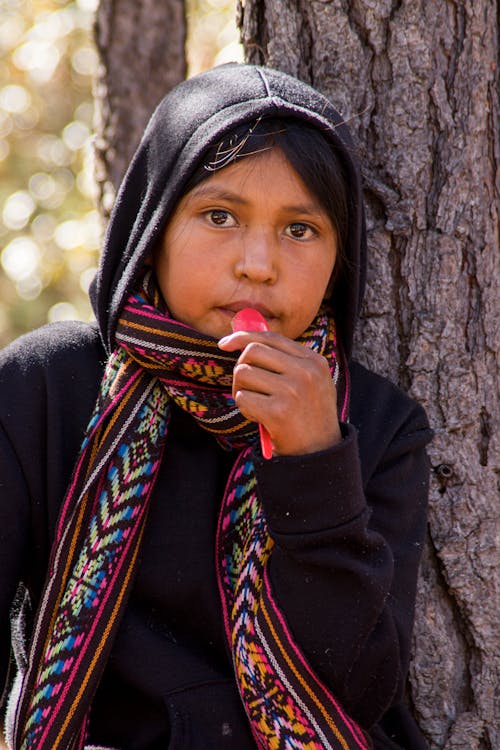 A young girl in a hoodie and scarf is leaning against a tree