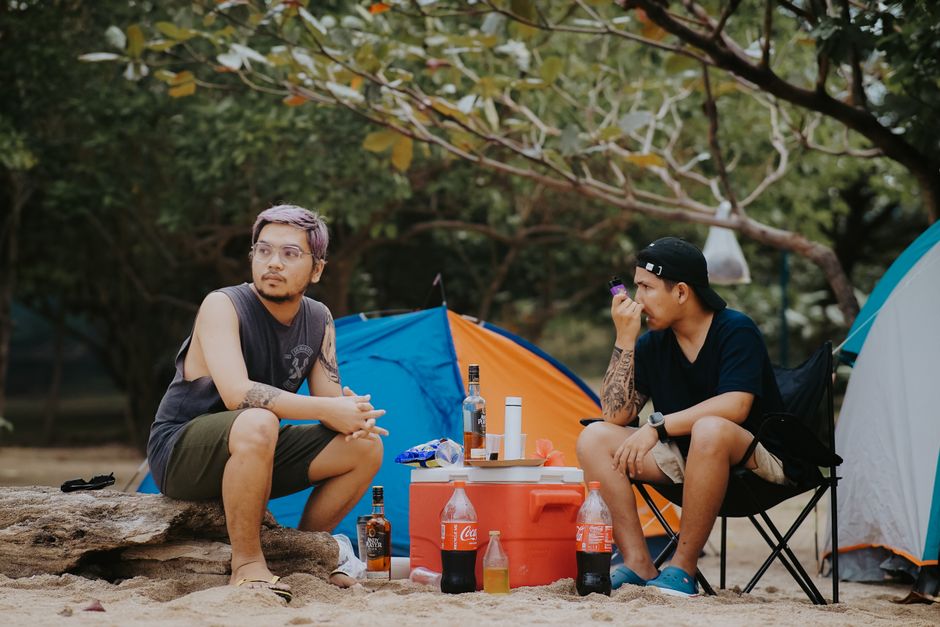 Two men enjoying a camping experience on the beach with drinks and snacks during summer.