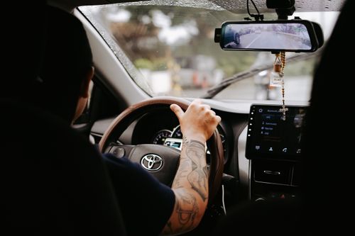 Free A driver's tattooed arm on the steering wheel inside a Toyota car during the day. Stock Photo