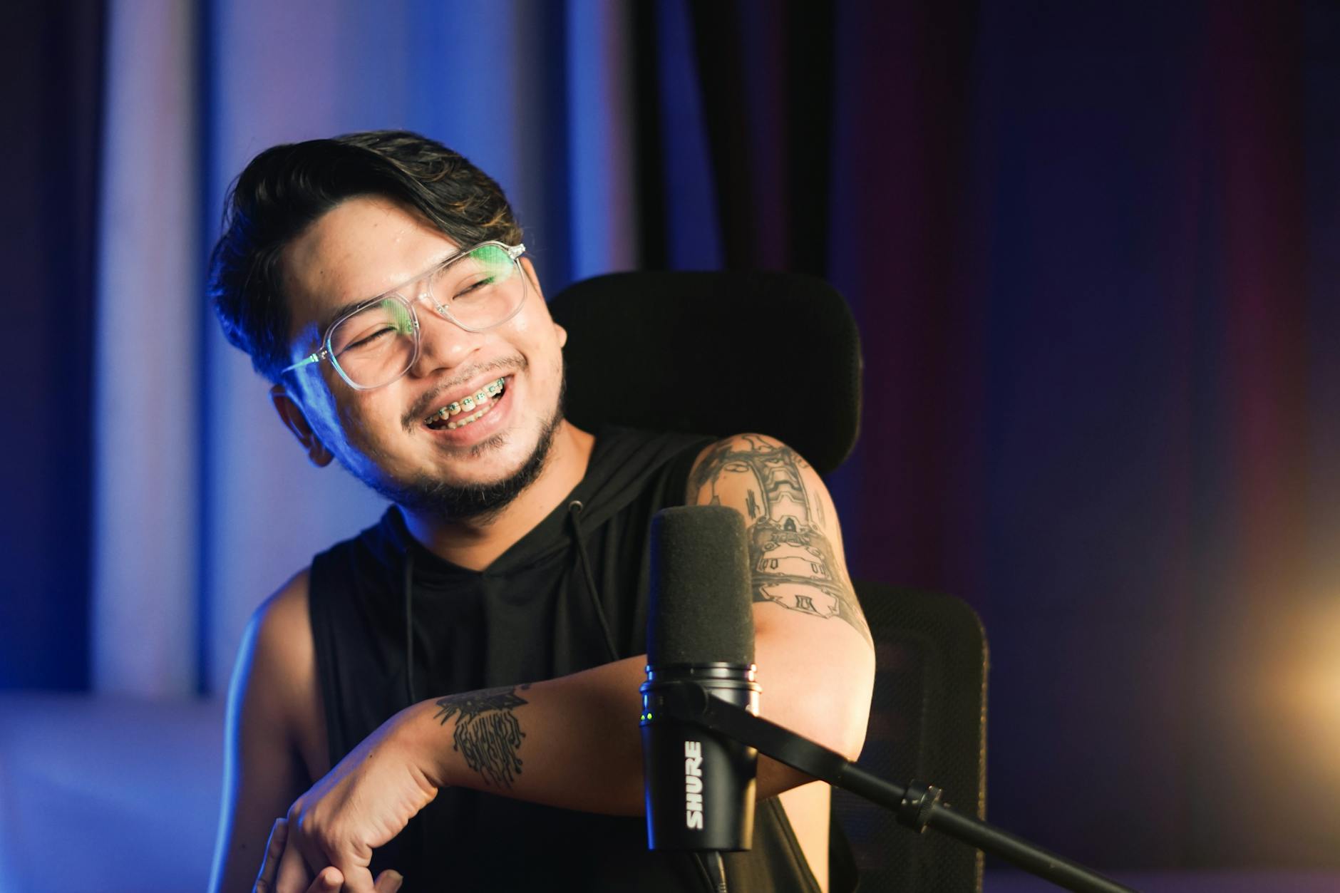 Young content creator smiling with microphone, tattoos, and eyeglasses in an indoor setting.