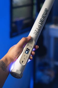 Close-up of a hand holding a battery-powered Amaran PT2C tube light in a dimly lit blue room.