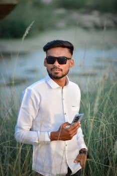 A fashionable man wearing sunglasses holds a smartphone in a grassy field, showcasing a cool and modern style.