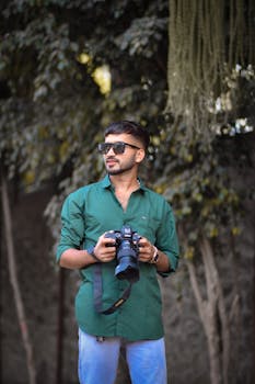 Young man in sunglasses holding a camera against a natural backdrop, showcasing modern fashion and photography.
