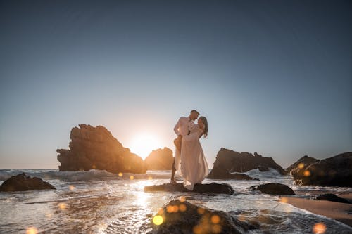 Woman in White Dress and Man in Shirt Hugging on Rocks on Sea Shore at Sunset