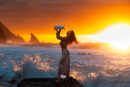 Woman in Dress Posing with Vase on Sea Shore under Yellow Sky at Sunset