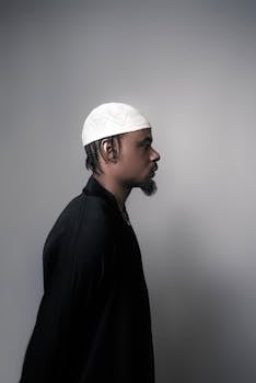 Profile portrait of a man in a black outfit and white cap, set against a neutral background.