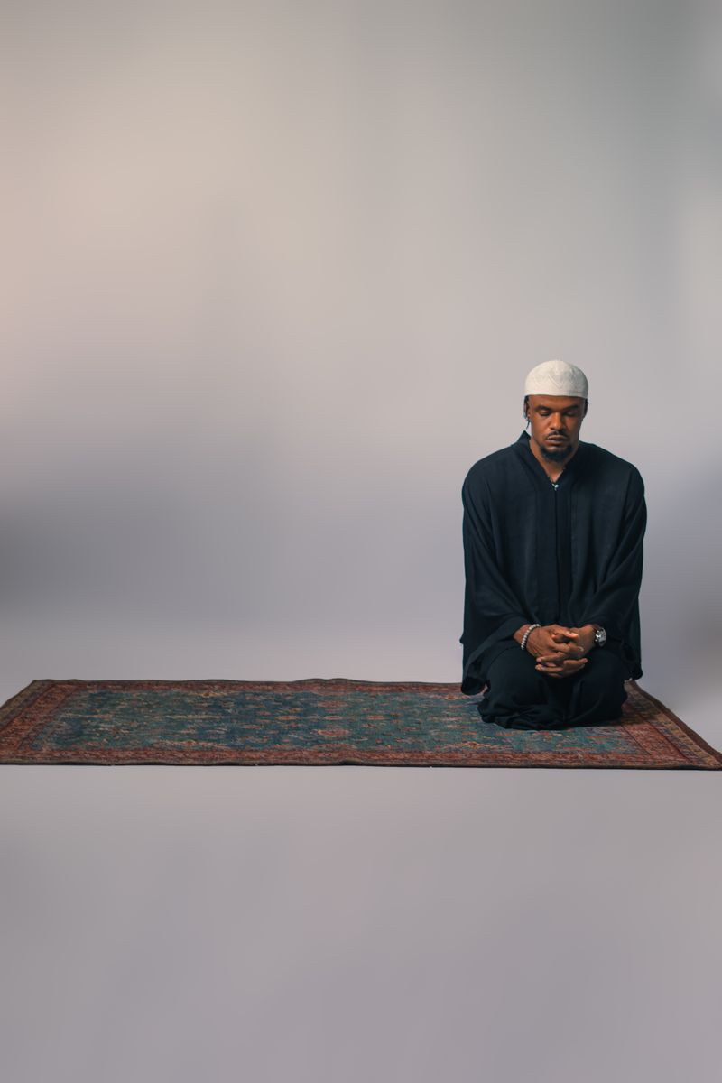 A man in traditional attire prays on a rug in a minimalist setting.