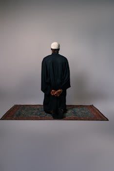 A man in a black robe meditates silently on a prayer rug in a minimalist setting.