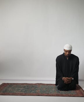 Adult man in black robe meditating on a prayer rug indoors, exuding calmness and spiritual focus.