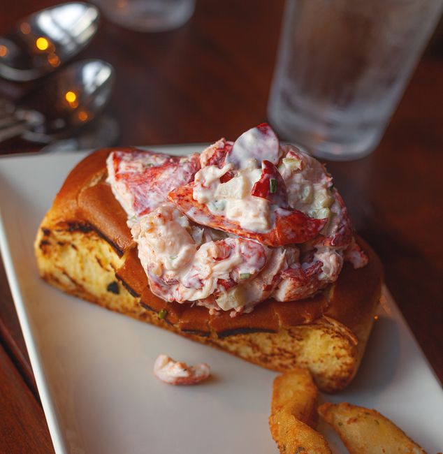A split-top bun toasted golden and stuffed with chunks of cold lobster meat lightly dressed in mayonnaise, topped with chopped chives, on a white plate with a lemon wedge.