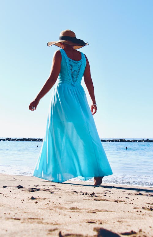 A woman in a blue dress and hat walking on the beach