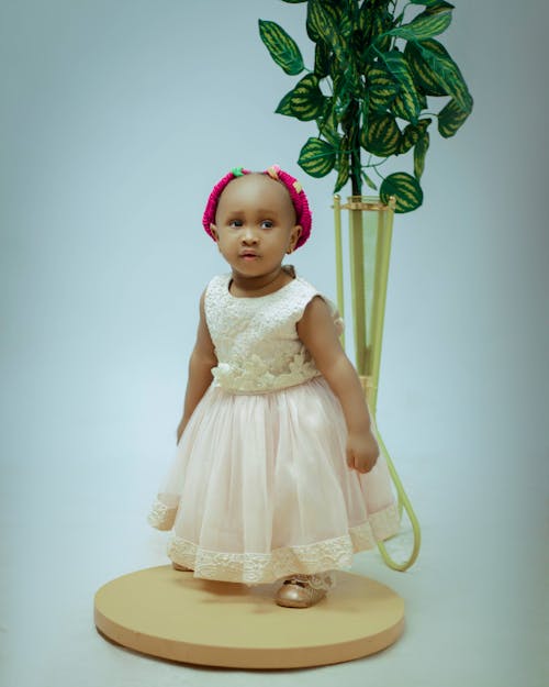 A baby girl in a pink dress standing next to a plant