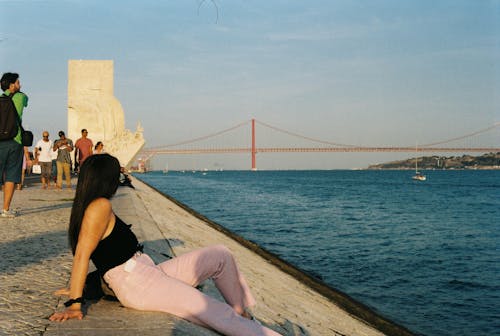 A woman sitting on the edge of a pier with a bridge in the background