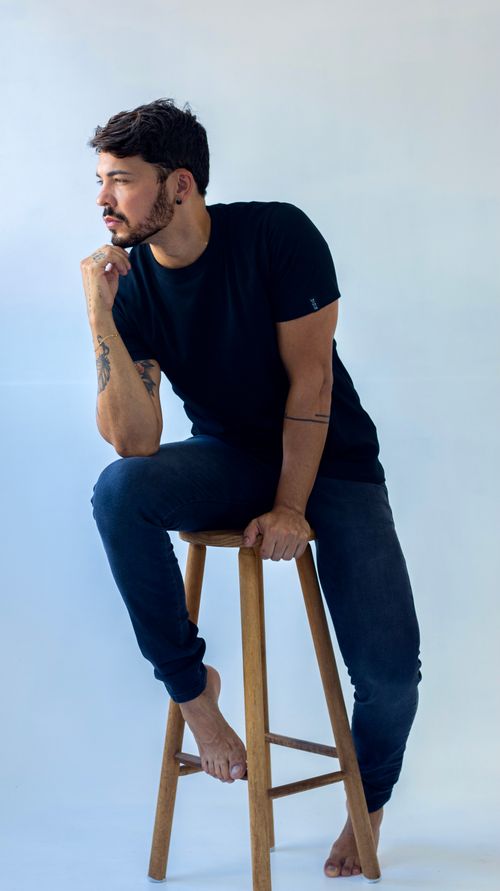 Free A man in a black t-shirt and jeans poses thoughtfully on a stool in studio setting. Stock Photo
