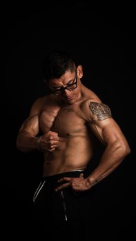 Portrait of a muscular man with tattoos and eyeglasses, standing against a black background, showcasing strength.