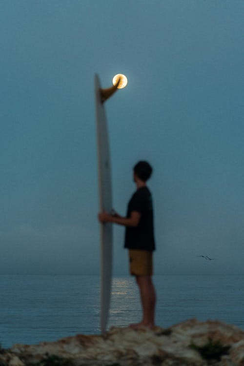 A man stands on the shore with a surfboard in the moonlight