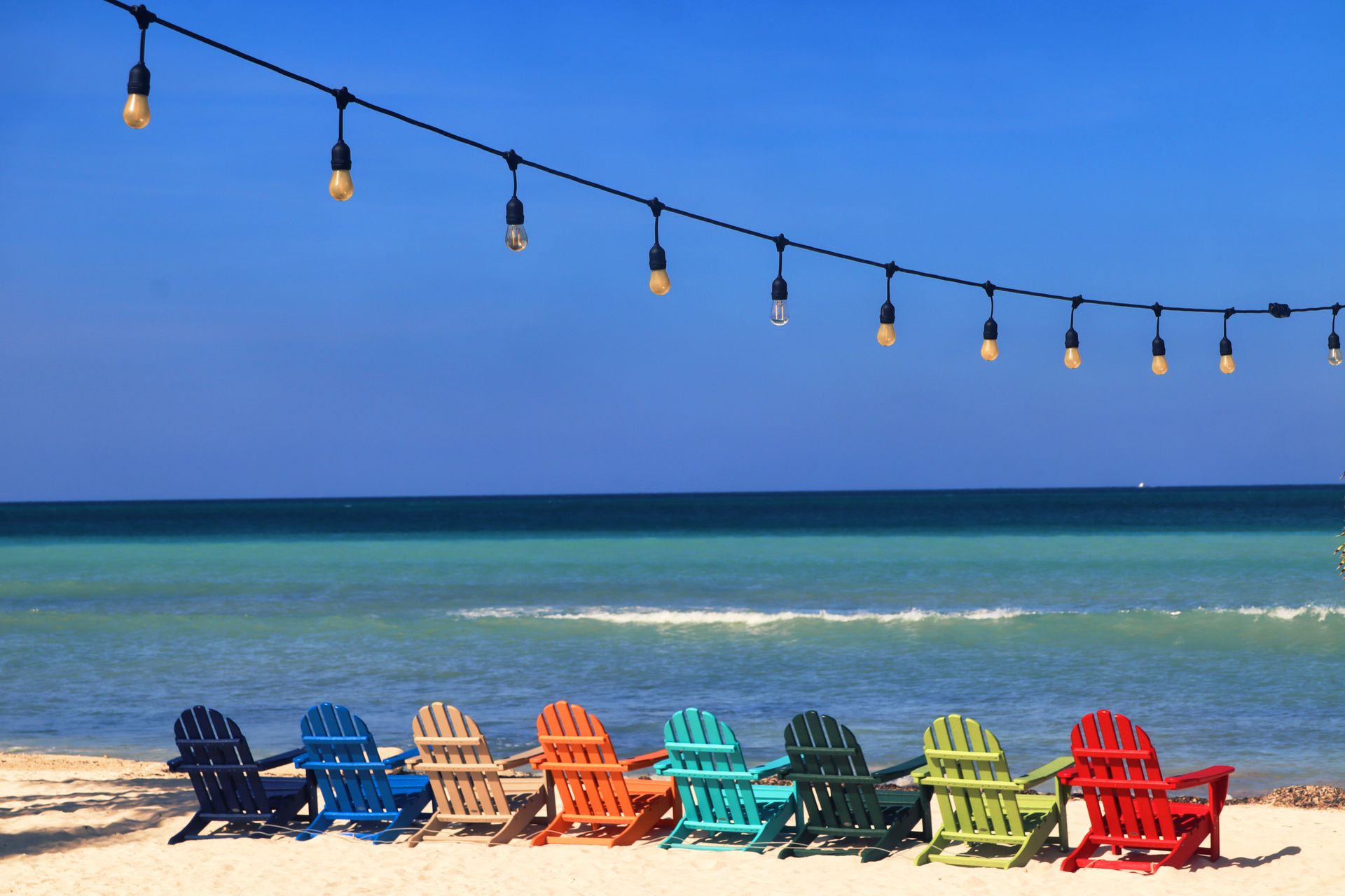 Colorful chairs face the ocean on a sunny Aruba shore under hanging lights.