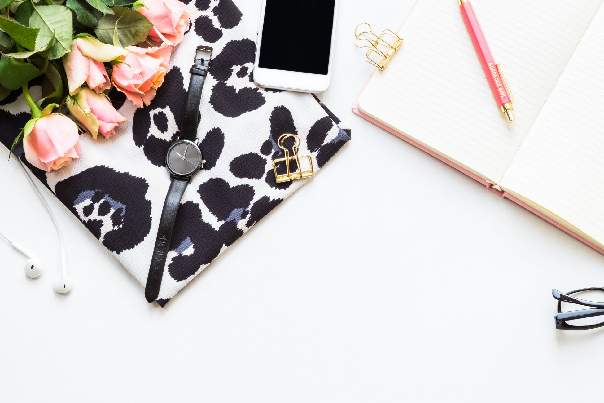 Stylish flat lay of office essentials including pink roses, watch, notebook, and accessories on white background.