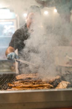 Chef grilling meat with tongs, surrounded by smoke in an indoor kitchen environment.