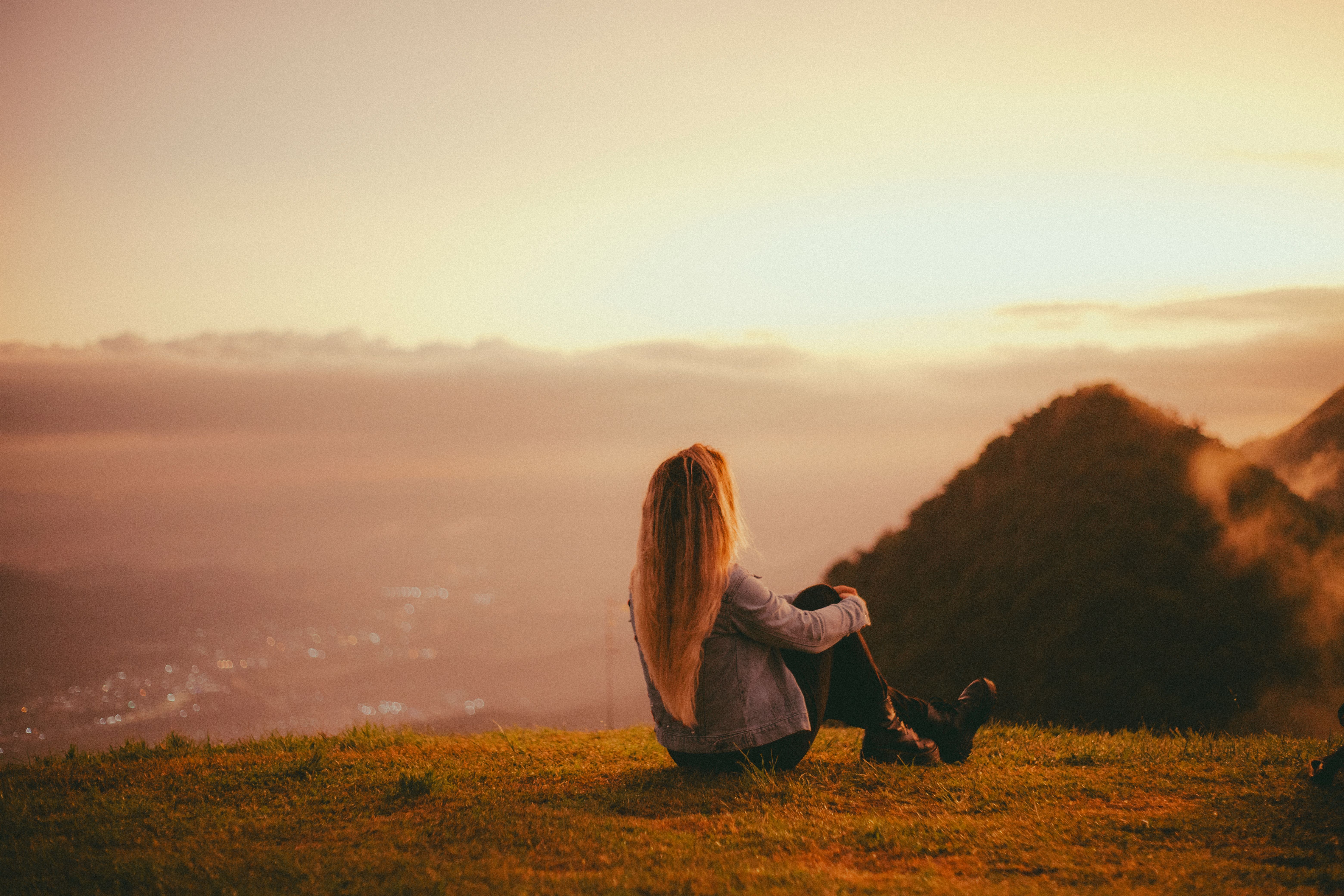 A woman with long hair relaxes on a hilltop, enjoying a tranquil sunset view.