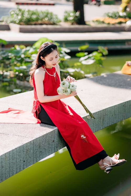 Woman in Traditional Clothing Sitting on Pier on Lake and Holding Flowers