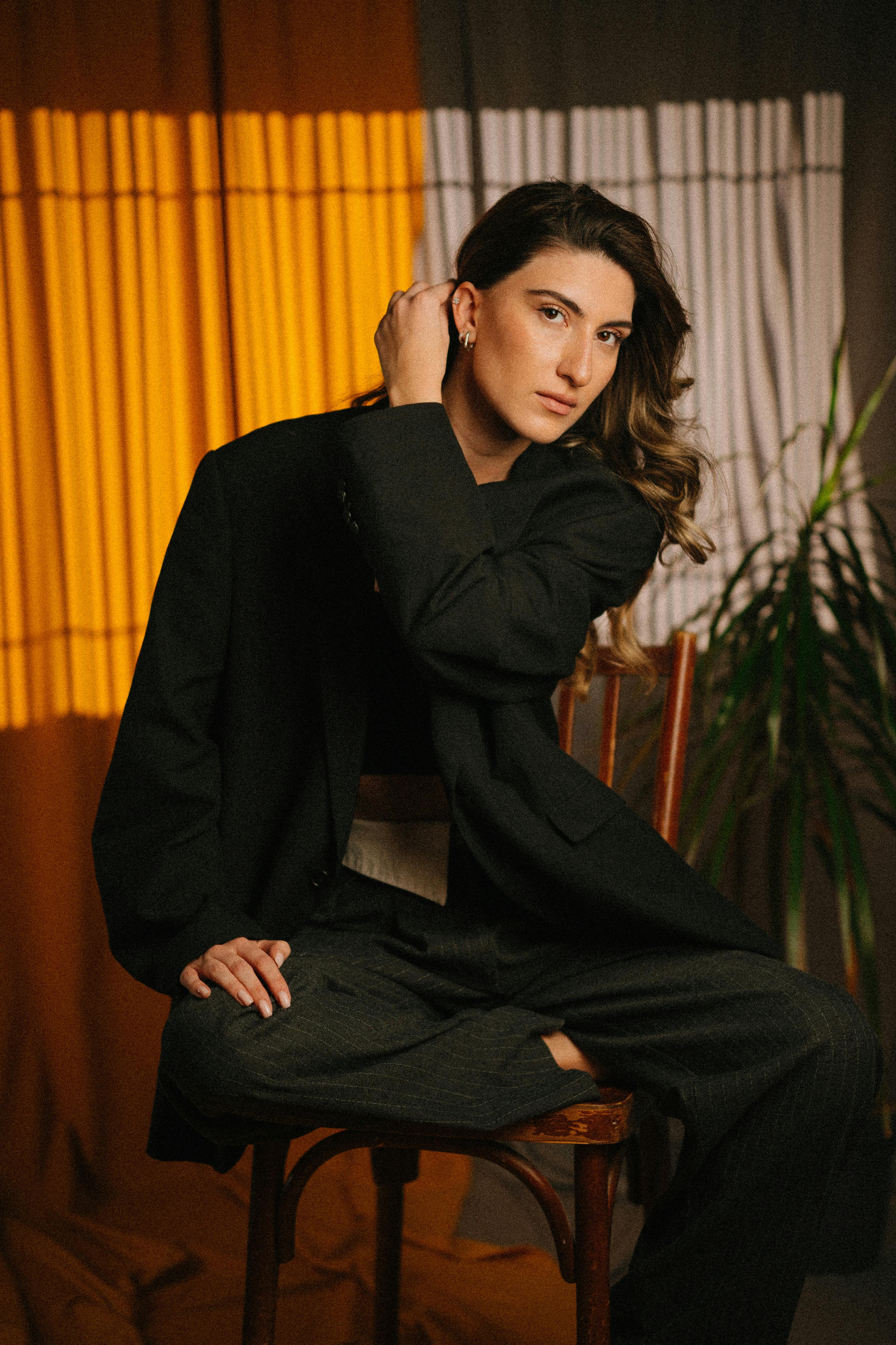 A stylish woman in a black suit poses gracefully on a chair with soft lighting and elegant composition.