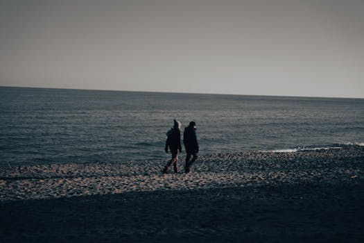 A couple in winter jackets stroll along a beach during dusk, highlighting a serene seaside scene.