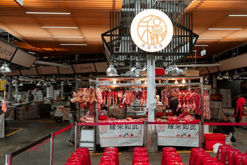 Free Active Asian market scene showcasing an indoor butcher stall with various meat cuts and vibrant red seating. Stock Photo