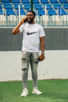 African man in casual wear talking on phone at a stadium with blue seats.