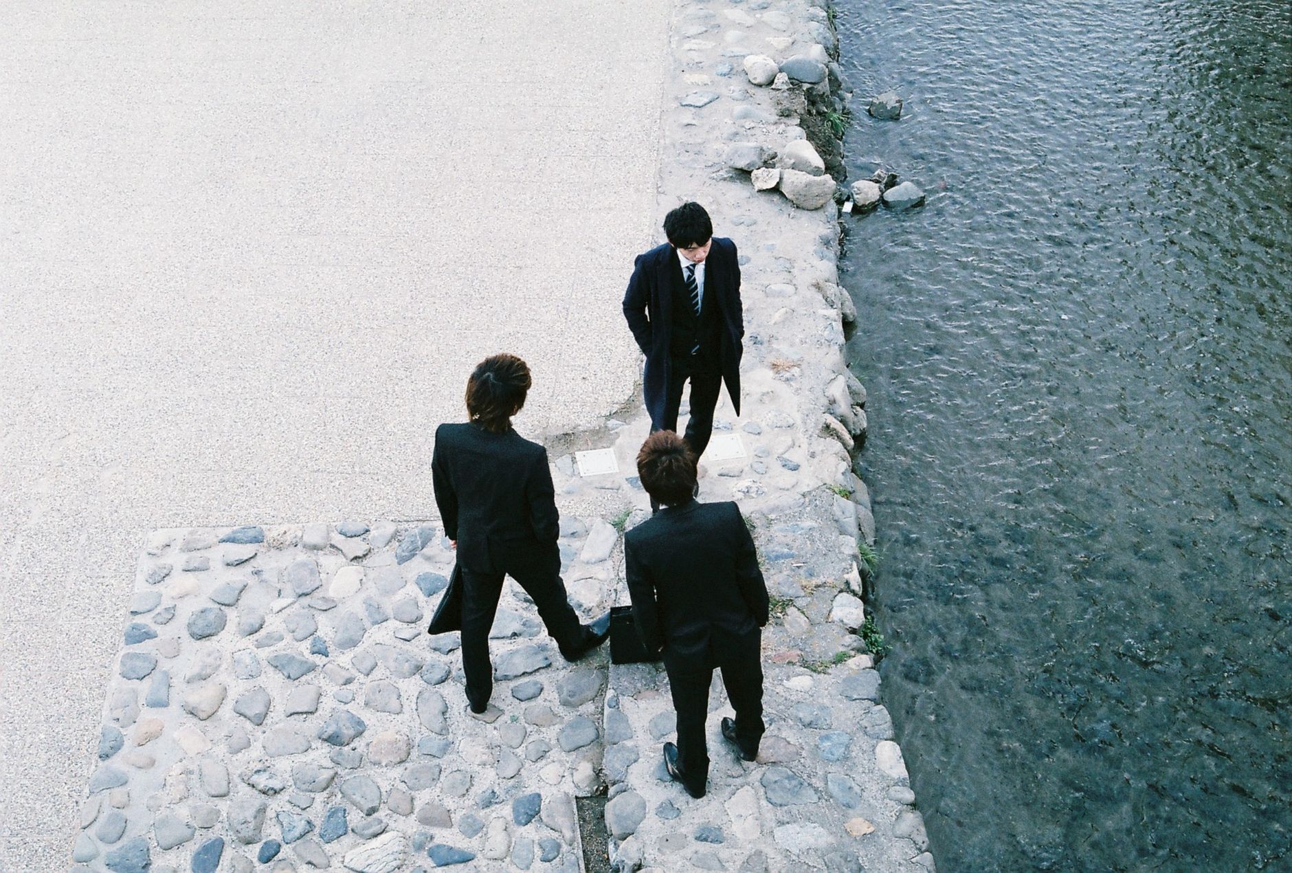 Three businessmen in suits stand by a river in Kyoto, Japan, engaged in discussion.