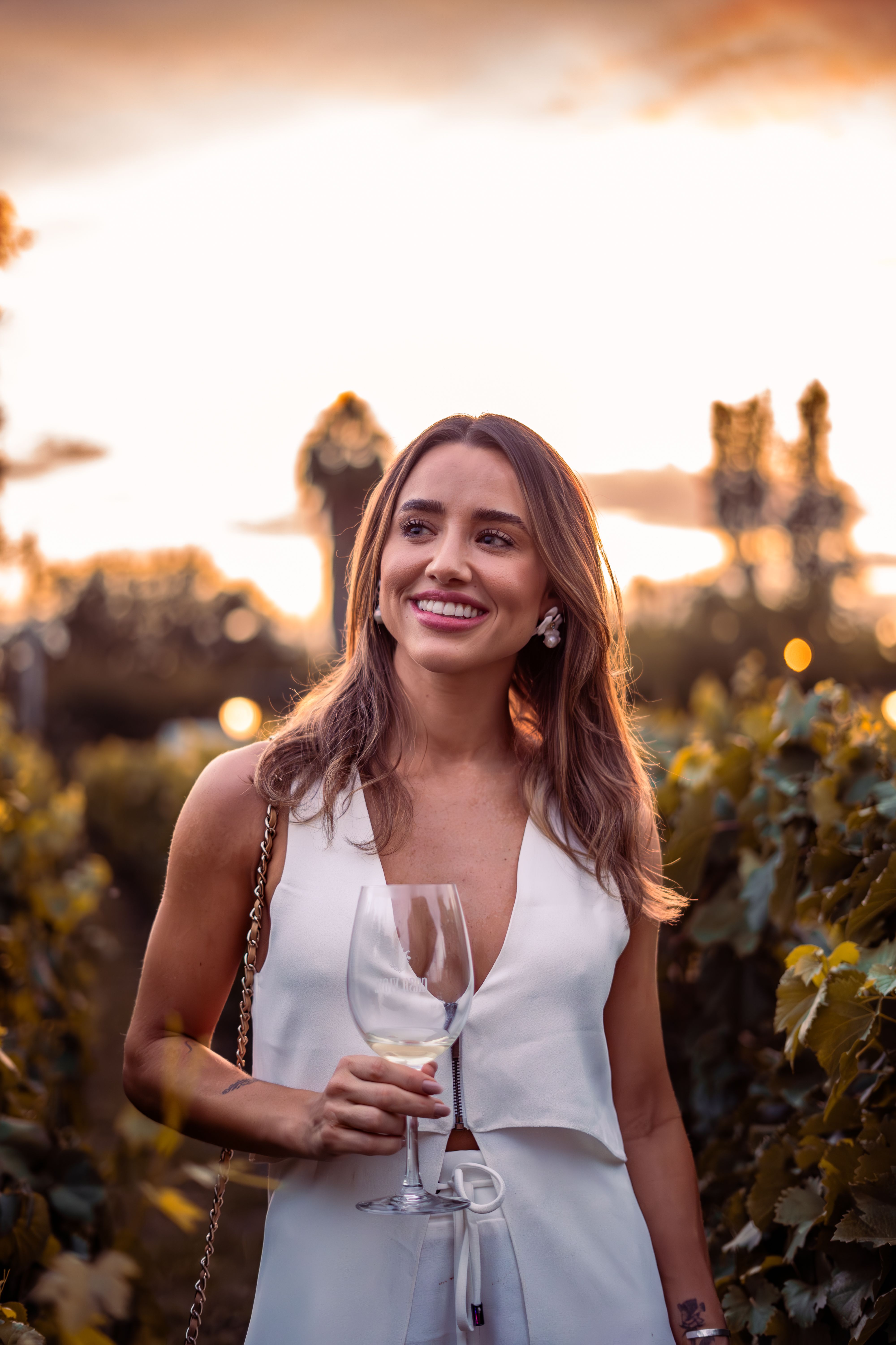 A woman with long brown hair smiles while holding a wine glass outdoors at sunset.