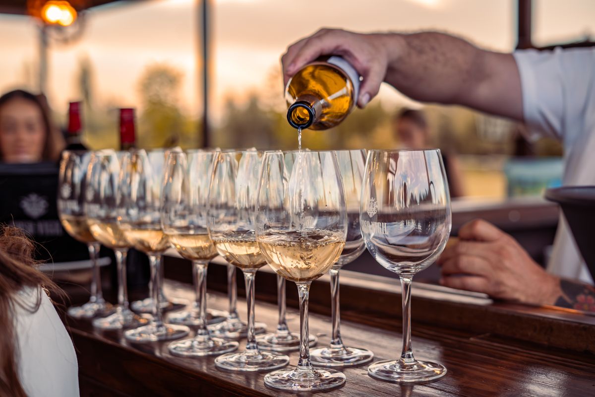 A waiter pouring white wine into a row of glasses on the bar at sunset