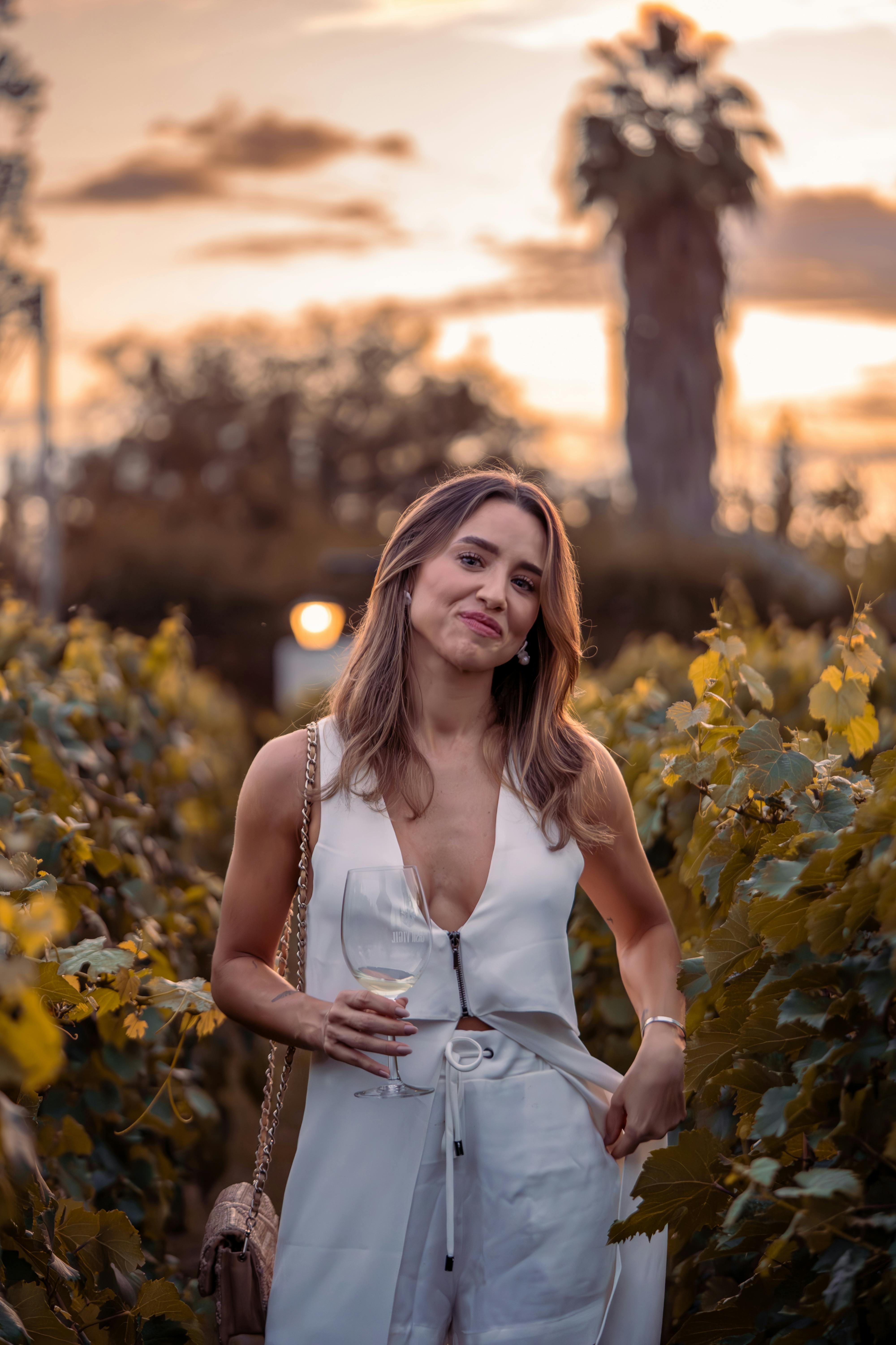 Charming woman with wine glass stands amidst lush vineyard at sunset.