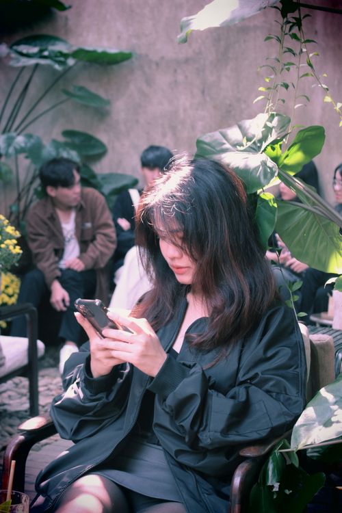 Free Asian woman in black jacket uses smartphone in a stylish indoor cafe with greenery. Stock Photo
