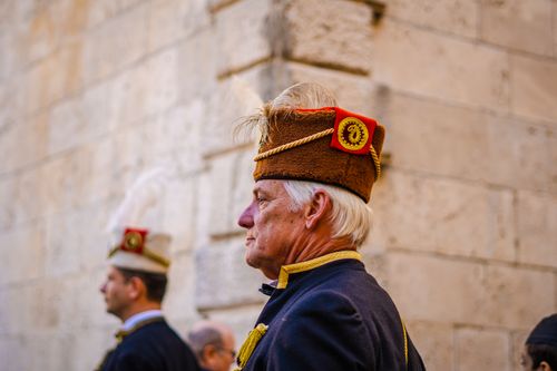 Free A senior man wearing traditional military attire stands against an urban stone wall. Stock Photo