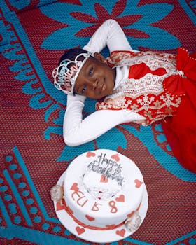 A joyful Nigerian girl in a festive dress celebrates her birthday with a decorated cake and crown.