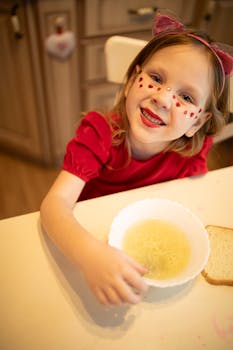 A young girl in cat ears and heart face paint smiles while eating soup indoors.