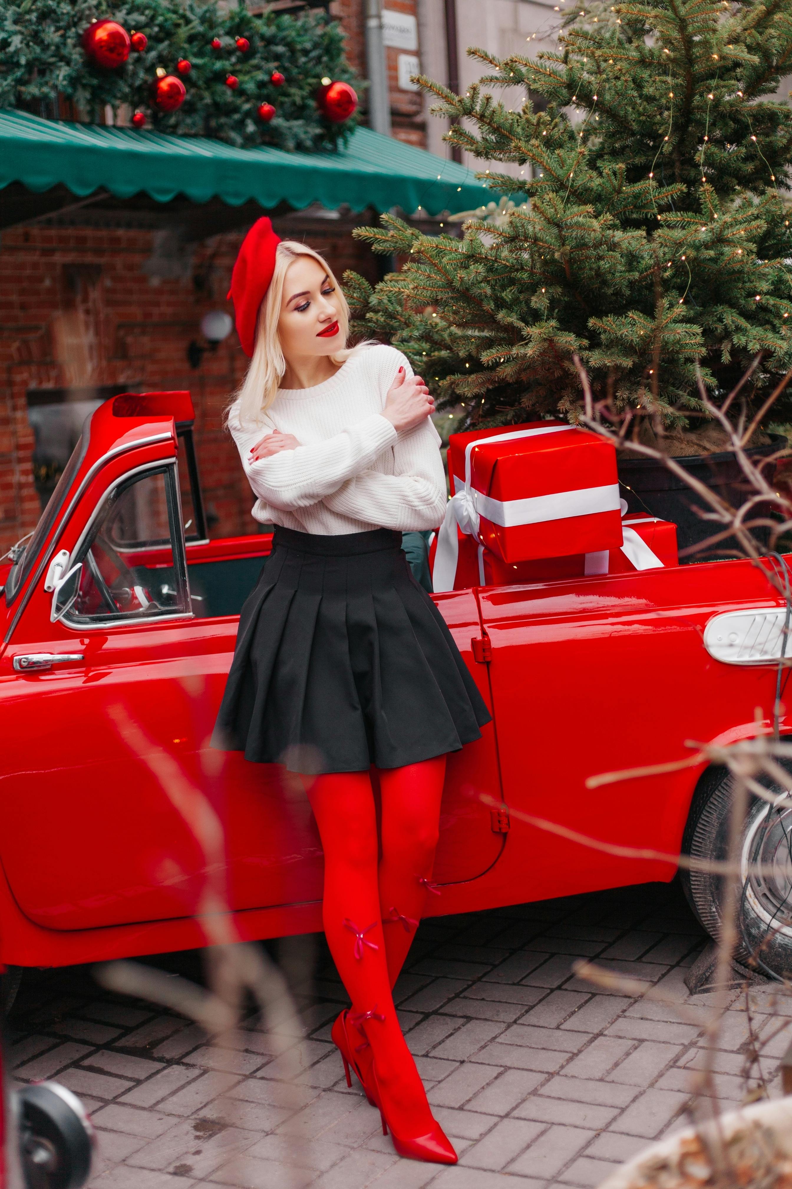 Woman in winter attire posing by red vintage car with Christmas gifts and tree.