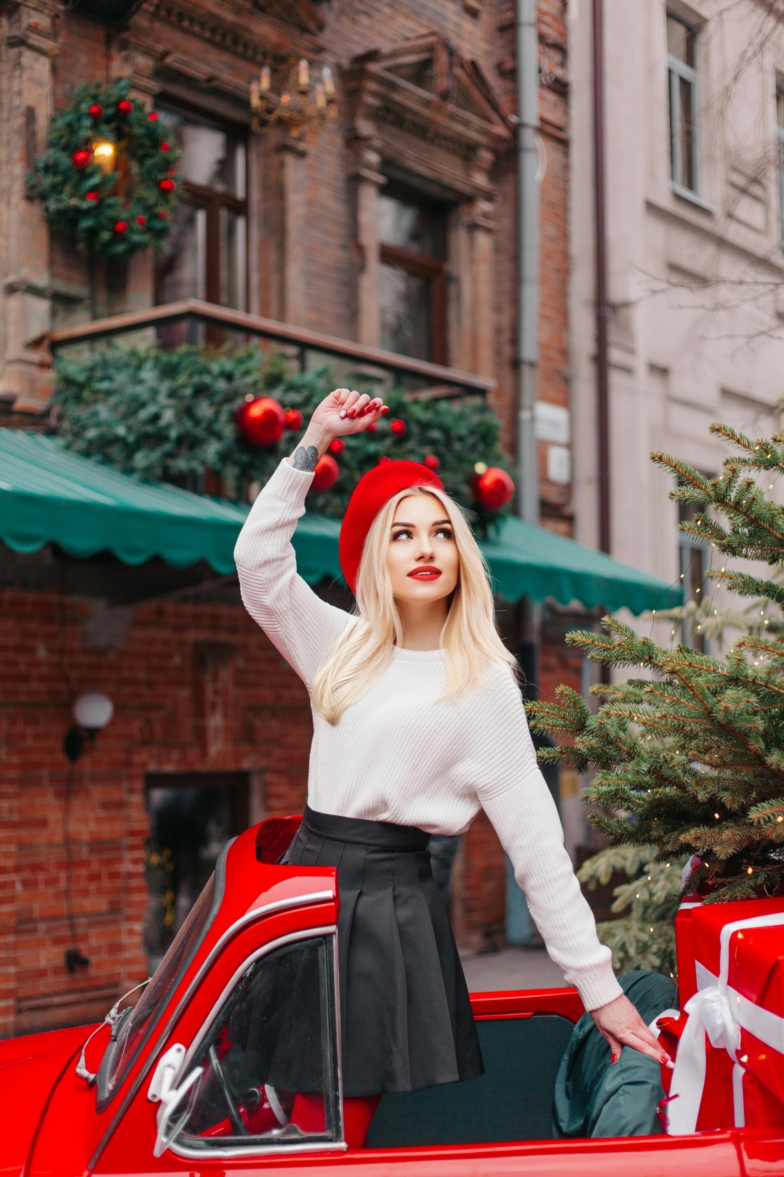 Stylish woman in red beret posing in a mini red car on a festive city street with Christmas decorations.