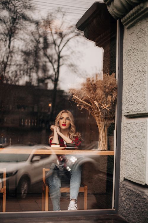 Free Young woman with red lips sitting in a café by the window, reflecting urban street view. Stock Photo