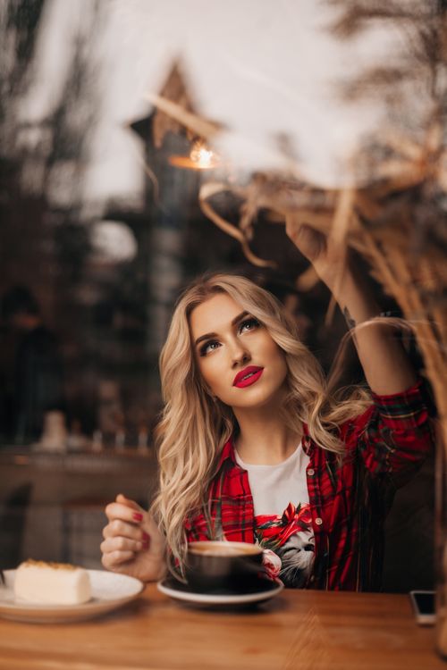 Free A blonde woman sits at a café table drinking coffee and enjoying a slice of cake, with a contemplative gaze outside the window. Stock Photo