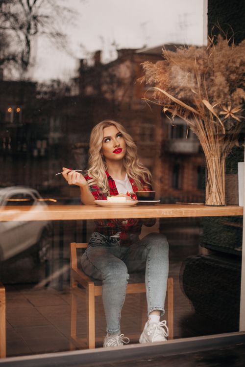Free Stylish woman in red shirt sipping coffee, seated in a modern café with a window view. Stock Photo
