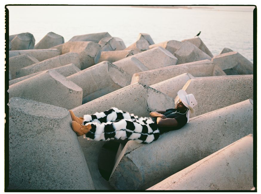 Fashionable man in bold checkered pants and cowboy hat relaxing on coastal concrete blocks at sunset.