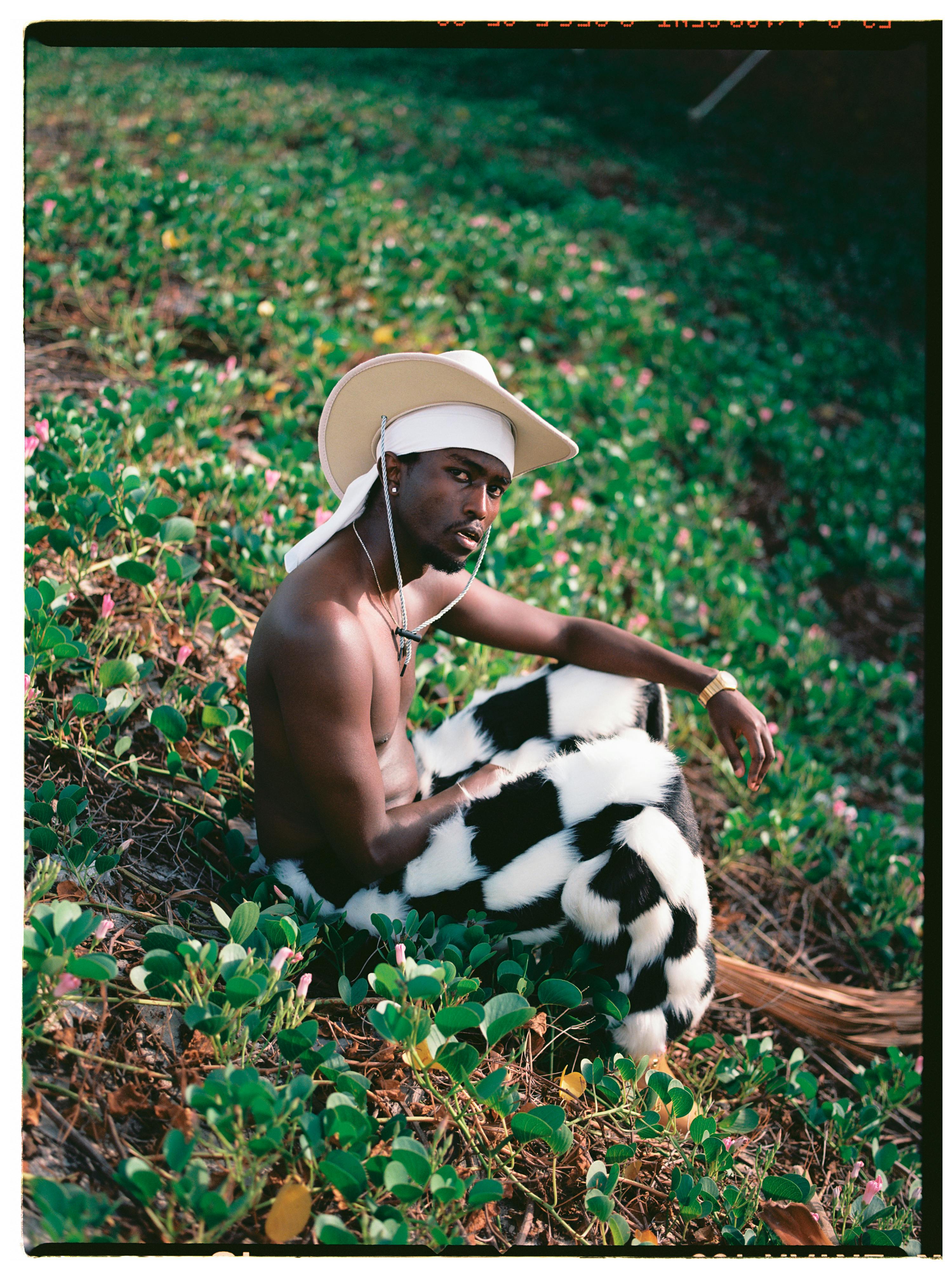 A man wearing a cowboy hat and bold patterned pants poses in a green outdoor setting.